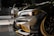 Close-up view of a customized car with a sleek design featuring carbon fiber and gold accents. In the background, another car is partially visible. The setting appears to be indoors with graffiti on the wall saying 'YOU'VE BEEN CLIMAX' and other details visible in the shadows.
