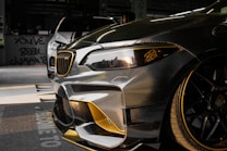 Close-up view of a customized car with a sleek design featuring carbon fiber and gold accents. In the background, another car is partially visible. The setting appears to be indoors with graffiti on the wall saying 'YOU'VE BEEN CLIMAX' and other details visible in the shadows.