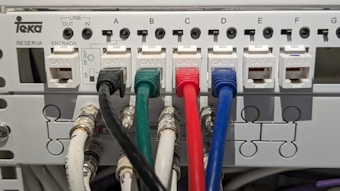 An electrical panel with multiple Category 6 (CAT 6) ethernet cables plugged into ports labeled from A to G. The cables are of different colors, including black, green, red, and blue, organized neatly into corresponding outlets. The panel includes labels for 'LINE OUT' and 'LINE IN' and the brand name 'Teka' is visible.