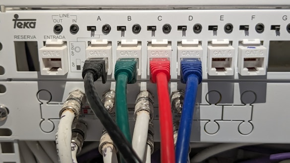 An electrical panel with multiple Category 6 (CAT 6) ethernet cables plugged into ports labeled from A to G. The cables are of different colors, including black, green, red, and blue, organized neatly into corresponding outlets. The panel includes labels for 'LINE OUT' and 'LINE IN' and the brand name 'Teka' is visible.