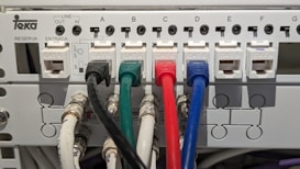 An electrical panel with multiple Category 6 (CAT 6) ethernet cables plugged into ports labeled from A to G. The cables are of different colors, including black, green, red, and blue, organized neatly into corresponding outlets. The panel includes labels for 'LINE OUT' and 'LINE IN' and the brand name 'Teka' is visible.