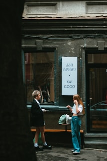 Two people are standing outside a building having a conversation. One person is wearing a white shirt and blue jeans, while the other is dressed in darker clothing. They are near a wall with a sign in both Georgian and English script that reads 'KAI GOGO'. A green bag is placed on a bench beside them.