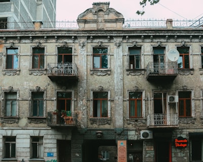 A weathered, vintage building with ornate architectural details, including decorative moldings and wrought iron balconies. The facade shows signs of aging with peeling paint and cracks. There are multiple windows with wooden frames, two balconies, and a satellite dish attached to the wall. Below, a neon sign reads 'Brocante Vintage' near a doorway.