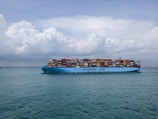 A large cargo ship with the text 'MAERSK LINE' printed on its side is sailing on a vast body of water. The ship is loaded with numerous colorful shipping containers stacked high. The sky above is partly cloudy with a mix of fluffy white and gray clouds.
