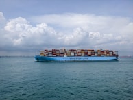 A large cargo ship with the text 'MAERSK LINE' printed on its side is sailing on a vast body of water. The ship is loaded with numerous colorful shipping containers stacked high. The sky above is partly cloudy with a mix of fluffy white and gray clouds.