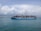 A large cargo ship with the text 'MAERSK LINE' printed on its side is sailing on a vast body of water. The ship is loaded with numerous colorful shipping containers stacked high. The sky above is partly cloudy with a mix of fluffy white and gray clouds.