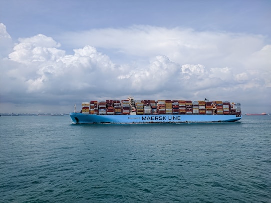 A large cargo ship with the text 'MAERSK LINE' printed on its side is sailing on a vast body of water. The ship is loaded with numerous colorful shipping containers stacked high. The sky above is partly cloudy with a mix of fluffy white and gray clouds.