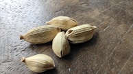 Several dried cardamom pods are scattered on a wooden surface. The pods have a distinctive ribbed texture and a beige color, with some signs of aging, such as slight browning at the tips.