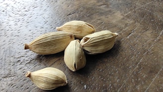 Several dried cardamom pods are scattered on a wooden surface. The pods have a distinctive ribbed texture and a beige color, with some signs of aging, such as slight browning at the tips.