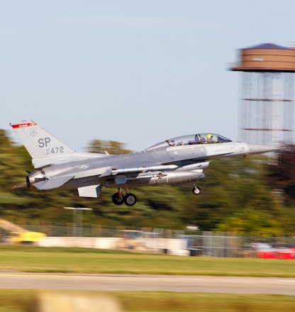 A sleek military fighter jet is taking off, with its wheels partially retracted, and is flying just above the runway. The background includes a blurry landscape of trees and a tall structure, indicating motion and speed.