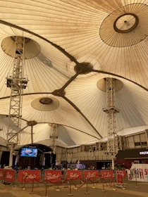 A large indoor venue with a high, tent-like ceiling supported by metal structures. The ceiling's fabric is pleated, creating a geometric pattern. Below, there are barricades with red banners and ropes, alongside an open area. A stage with electronic displays is visible, and the setting appears to be a public space.