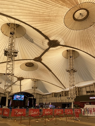 A large indoor venue with a high, tent-like ceiling supported by metal structures. The ceiling's fabric is pleated, creating a geometric pattern. Below, there are barricades with red banners and ropes, alongside an open area. A stage with electronic displays is visible, and the setting appears to be a public space.