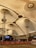 A large indoor venue with a high, tent-like ceiling supported by metal structures. The ceiling's fabric is pleated, creating a geometric pattern. Below, there are barricades with red banners and ropes, alongside an open area. A stage with electronic displays is visible, and the setting appears to be a public space.