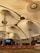 A large indoor venue with a high, tent-like ceiling supported by metal structures. The ceiling's fabric is pleated, creating a geometric pattern. Below, there are barricades with red banners and ropes, alongside an open area. A stage with electronic displays is visible, and the setting appears to be a public space.