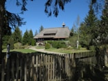 A rustic wooden house with a thatched roof is surrounded by lush greenery and tall trees. A picket fence encloses the yard, which is well-manicured with pathways and shrubs. The sky is clear and blue, enhancing the serene atmosphere.