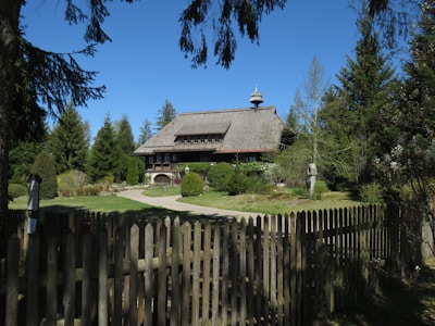 A rustic wooden house with a thatched roof is surrounded by lush greenery and tall trees. A picket fence encloses the yard, which is well-manicured with pathways and shrubs. The sky is clear and blue, enhancing the serene atmosphere.