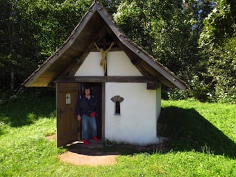 A small, rustic chapel with a wooden roof and white walls is nestled in a lush green area with dense trees. A figure of Jesus on a crucifix is mounted above the entrance. A person is standing in the doorway, partially in shadow due to the bright sunlight.