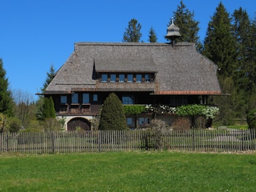 A rustic wooden house with a steep, shingled roof is set in a lush landscape. Large windows are symmetrically aligned on the upper level, and the lower level is adorned with greenery and shrubs. A wooden fence surrounds the property, and tall pine trees are visible in the background under a clear blue sky.