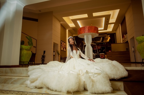 A person in an elegant white gown, embellished with lace, sits gracefully in a grand interior space. The setting features modern architecture with high ceilings and stylish lighting fixtures. Large green plants in decorative pots are placed around the area, adding a touch of nature to the sophisticated environment. A prominent red and white sculpture or lamp is centrally located behind the person, contributing to the artistic ambiance.