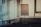 An empty conference room with vertical blinds partially open, allowing light to filter through. In the center, a blank flipchart stands on a mobile easel. A table is visible in the corner, holding a coiled cable and a pair of glasses.