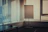 An empty conference room with vertical blinds partially open, allowing light to filter through. In the center, a blank flipchart stands on a mobile easel. A table is visible in the corner, holding a coiled cable and a pair of glasses.