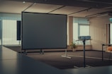 An empty conference room featuring a large projection screen and a projector mounted on a stand. The room is well-lit with natural light coming through the vertical blinds covering the windows. A potted plant is placed beside the screen, adding a touch of greenery to the neutral and modern interior. Tables and chairs are arranged around the room, suggesting a setup for presentations or meetings.