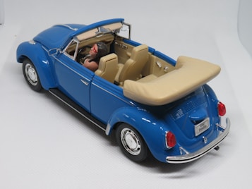 A miniature model of a classic convertible car with a bright blue exterior and tan interior. The car has detailed features including a license plate marked 'VW Beetle', chrome elements, and a driver figure wearing red sunglasses. The model is placed against a plain, light background.
