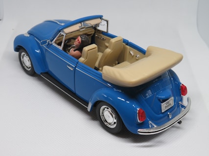 A miniature model of a classic convertible car with a bright blue exterior and tan interior. The car has detailed features including a license plate marked 'VW Beetle', chrome elements, and a driver figure wearing red sunglasses. The model is placed against a plain, light background.
