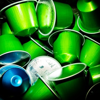A collection of coffee capsules, predominantly in bright green with one capsule in blue, stacked together. The metallic tops of some capsules display text and logos.