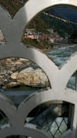 A scenic view of a river flowing through a mountainous region with buildings on the hillside. Colorful prayer flags are strung across the scene, and the foreground features an ornate metal railing.