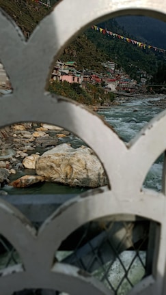 A scenic view of a river flowing through a mountainous region with buildings on the hillside. Colorful prayer flags are strung across the scene, and the foreground features an ornate metal railing.