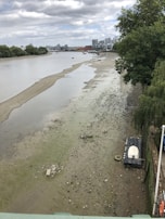 A river with very low water levels exposes a muddy and debris-filled riverbed. There is a small white boat resting on a platform near the shoreline. The surrounding area includes a tree-lined bank and distant residential buildings under a cloudy sky.