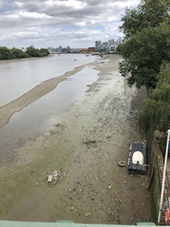 A river with very low water levels exposes a muddy and debris-filled riverbed. There is a small white boat resting on a platform near the shoreline. The surrounding area includes a tree-lined bank and distant residential buildings under a cloudy sky.
