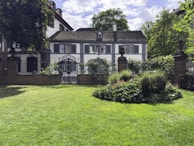 A picturesque garden scene featuring a well-maintained lawn and a circular flower bed surrounded by lush greenery. In the background, there is a charming white house with dark shutters and a slate roof, partly obscured by trees and framed by a wrought iron fence and gate.