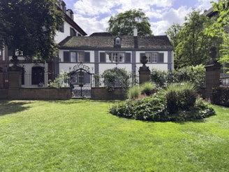 A picturesque garden scene featuring a well-maintained lawn and a circular flower bed surrounded by lush greenery. In the background, there is a charming white house with dark shutters and a slate roof, partly obscured by trees and framed by a wrought iron fence and gate.