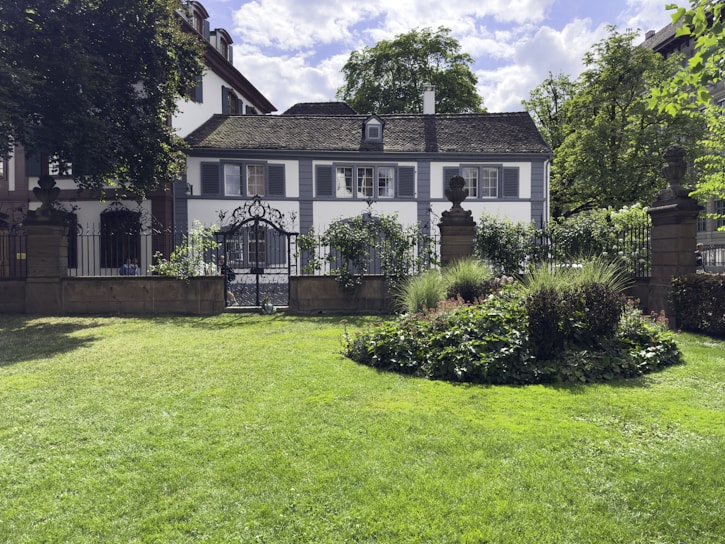 A picturesque garden scene featuring a well-maintained lawn and a circular flower bed surrounded by lush greenery. In the background, there is a charming white house with dark shutters and a slate roof, partly obscured by trees and framed by a wrought iron fence and gate.