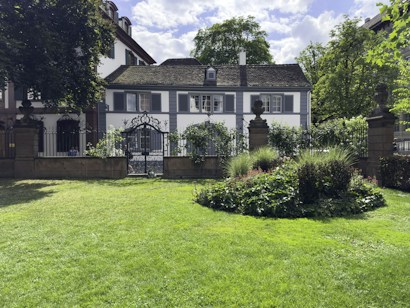 A picturesque garden scene featuring a well-maintained lawn and a circular flower bed surrounded by lush greenery. In the background, there is a charming white house with dark shutters and a slate roof, partly obscured by trees and framed by a wrought iron fence and gate.
