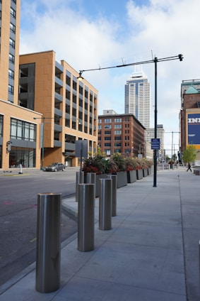 An urban street scene featuring a sidewalk lined with tall cylindrical metal bollards and large rectangular planters containing decorative plants. In the background, modern mid-rise buildings in shades of beige and brown are prominent, with a taller skyscraper further in the distance. The sky is partly cloudy, and the street seems quiet with minimal vehicular activity.