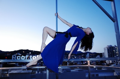 A woman in a vibrant blue dress is suspended horizontally in mid-air against a cityscape backdrop. She is holding onto a vertical pole with one hand, her hair flowing downward. The sky is transitioning between daylight and dusk, with buildings and the word 'rooftop' visible in the background.