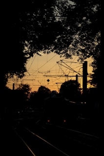 Cables and electrical wires form intricate patterns against a sunset sky, with silhouetted trees framing the scene. A dimly lit train track runs through the middle of the image.