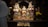 A colorful and ornate display of traditional wooden deities in a richly decorated shrine with floral arrangements. The figures are adorned with vibrant clothing and intricate headpieces. A person in a white garment performs a ritual with a lit candle, suggesting a spiritual or religious ceremony.