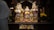 A colorful and ornate display of traditional wooden deities in a richly decorated shrine with floral arrangements. The figures are adorned with vibrant clothing and intricate headpieces. A person in a white garment performs a ritual with a lit candle, suggesting a spiritual or religious ceremony.
