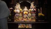 A colorful and ornate display of traditional wooden deities in a richly decorated shrine with floral arrangements. The figures are adorned with vibrant clothing and intricate headpieces. A person in a white garment performs a ritual with a lit candle, suggesting a spiritual or religious ceremony.