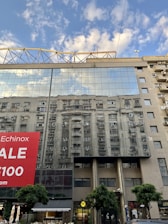 The glass facade of a modern building reflects the image of another building, creating a mirrored effect. A large red sale banner is prominently displayed on the left side. The sky in the background is partly cloudy, enhancing the reflective quality of the glass. Trees and various small shops are visible at street level.