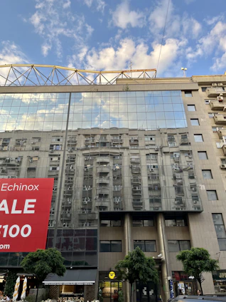 The glass facade of a modern building reflects the image of another building, creating a mirrored effect. A large red sale banner is prominently displayed on the left side. The sky in the background is partly cloudy, enhancing the reflective quality of the glass. Trees and various small shops are visible at street level.