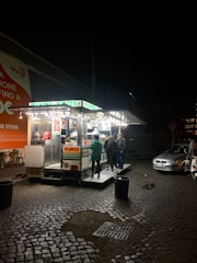 A small food stand is illuminated against the night sky, with bright lights showcasing a menu and various food items. Several people are gathered around, engaging with the vendor. The stand is set on a cobblestone street with a couple of parked cars nearby. An orange advertisement is visible on the wall to the left, and a few chairs are set up outside.