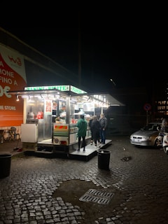 A small food stand is illuminated against the night sky, with bright lights showcasing a menu and various food items. Several people are gathered around, engaging with the vendor. The stand is set on a cobblestone street with a couple of parked cars nearby. An orange advertisement is visible on the wall to the left, and a few chairs are set up outside.