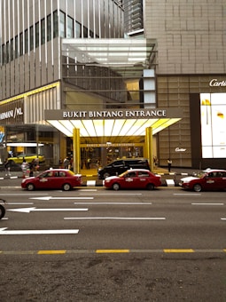 The image captures the entrance of Bukit Bintang, characterized by a large, illuminated canopy. Several red taxis are parked in front of the entrance, with people walking nearby. A tall, modern building with reflective glass windows forms the backdrop, while brand logos are visible on the sides of the building.