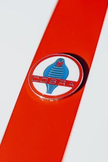 A close-up view of an emblem featuring a stylized blue cobra on a white circle with a red background. The word 'COBRA' is written below the snake in bold, red letters. The emblem is set against a bright orange stripe that contrasts with the white base color.