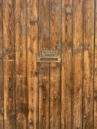 A wooden door with a textured surface, featuring vertical planks and a small metal sign. The sign reads 'Non si accetta pubblicità' above a metal letter slot inscribed with 'Lettere'. The wood has a natural, rustic appearance with visible grain patterns and knots.